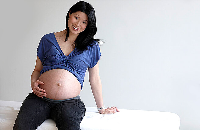 A pregnant Asian woman is sitting on the medical bad and she is holding her belly with her hand. 